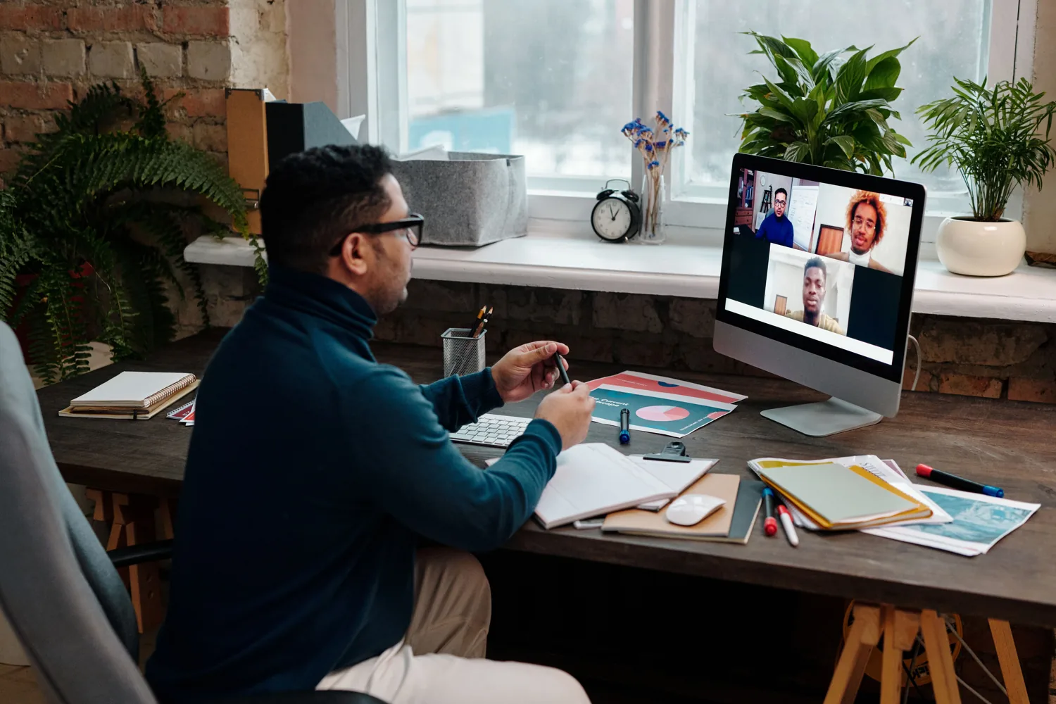 A man on a video call at his desk, with four colleagues visible on the screen, as an AI note-taker records the meeting.