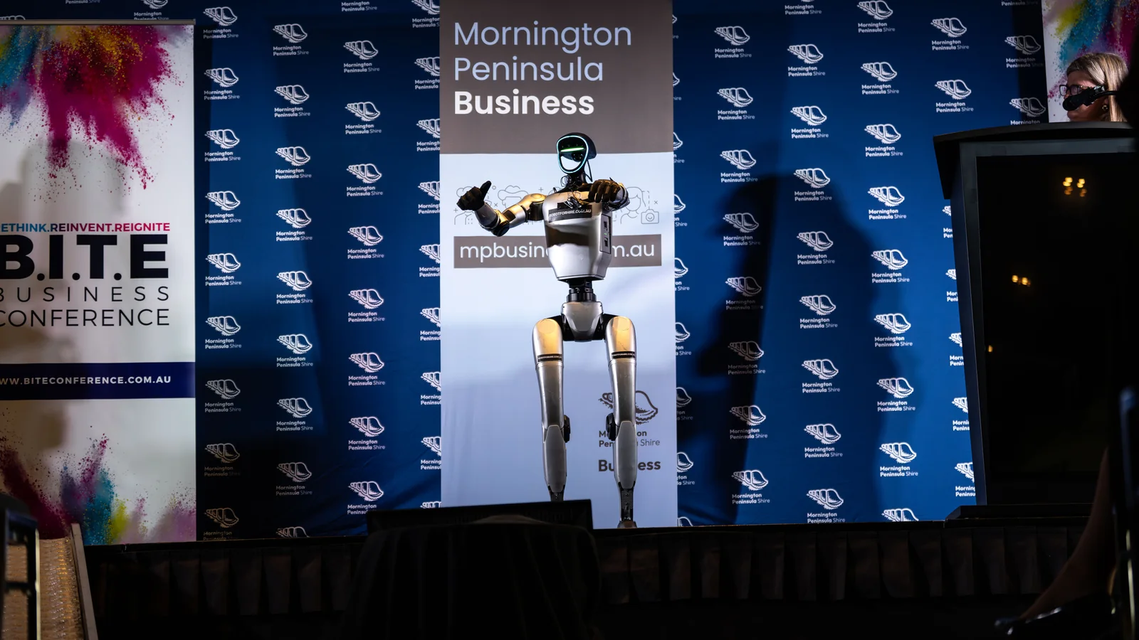 A humanoid robot gesturing on stage at the B.I.T.E Business Conference 2026 in front of the Mornington Peninsula Business backdrop, the kind of AI that draws a crowd while the real risk arrives by email.