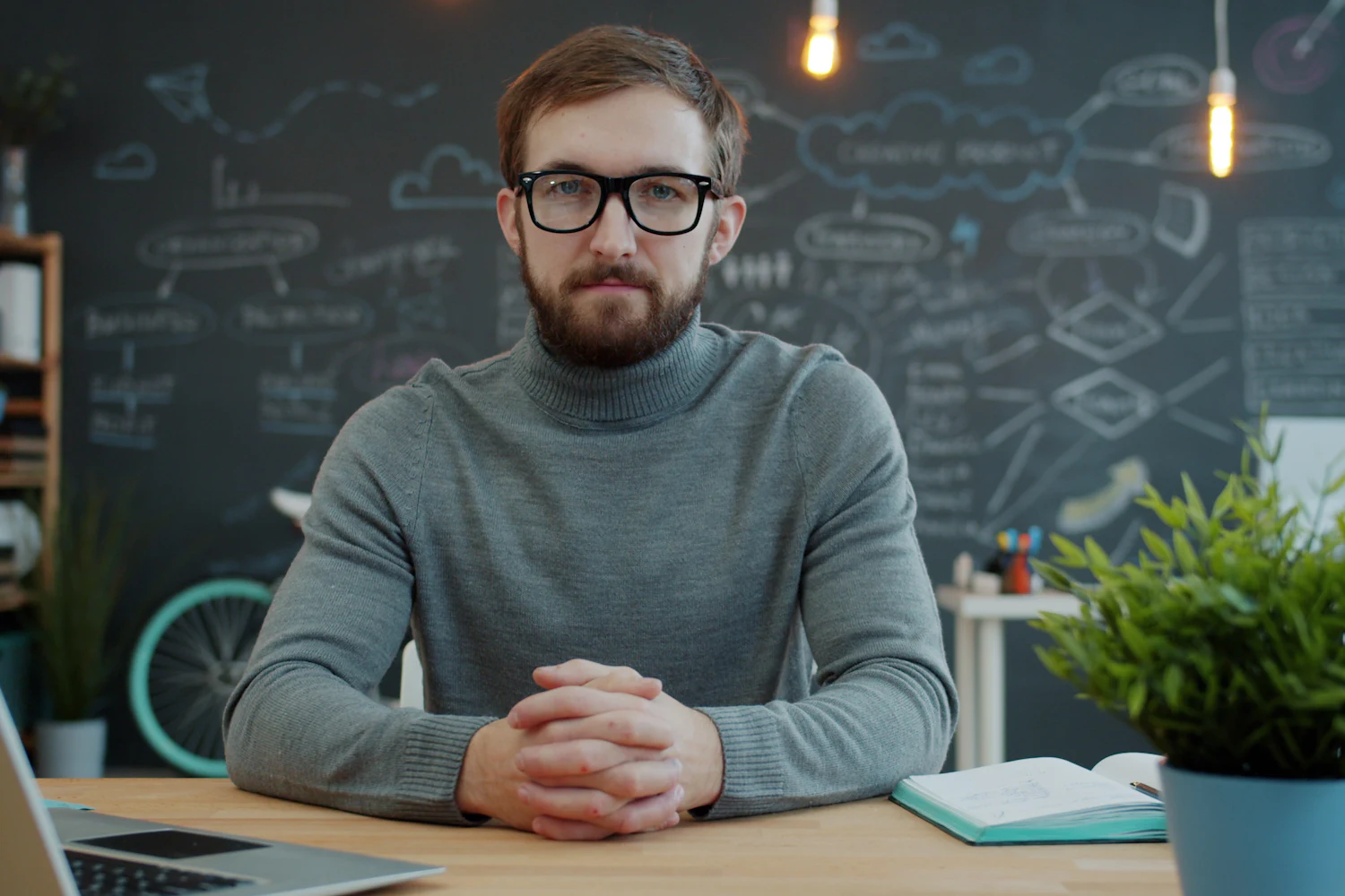 A business owner sitting at a desk, hands clasped, thinking through a decision, with strategy diagrams on the wall behind.