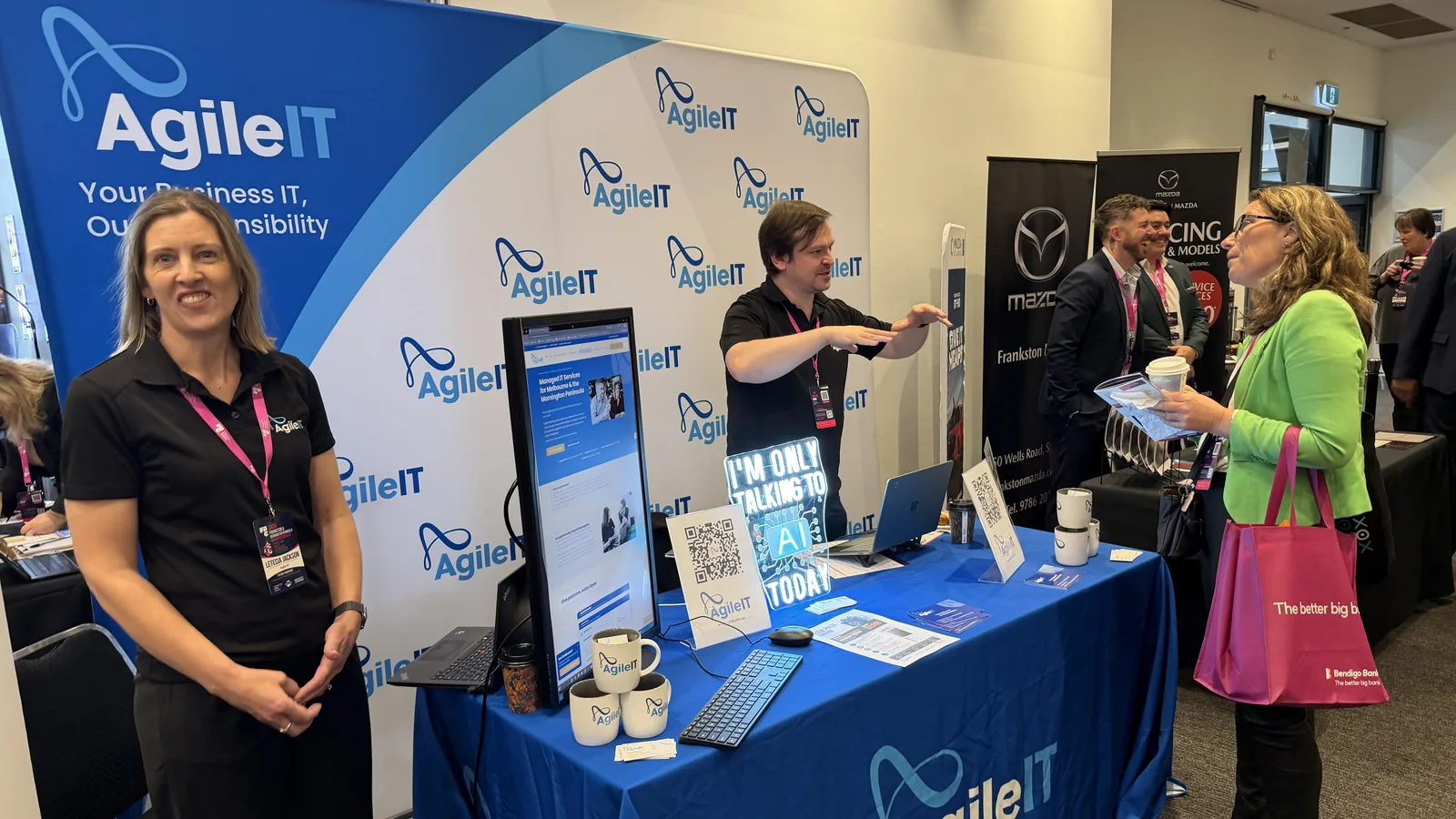 The Agile IT stand at the Frankston Business Festival 2026, with the branded backdrop, the I'm only talking to AI today neon sign, and the team talking with visitors.