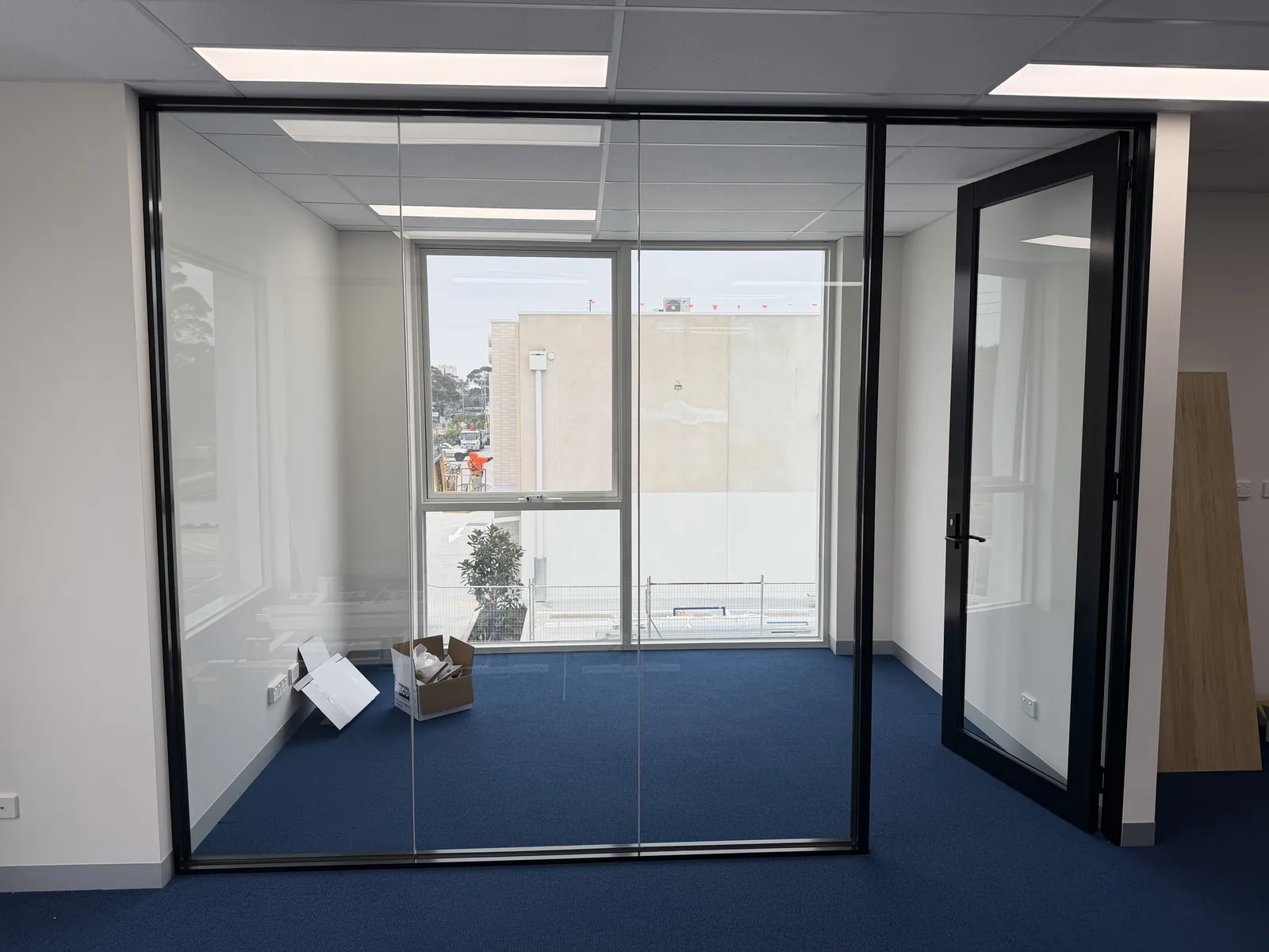 Another glass-partitioned office upstairs, black-framed glass with a window out to the neighbouring building, blue carpet