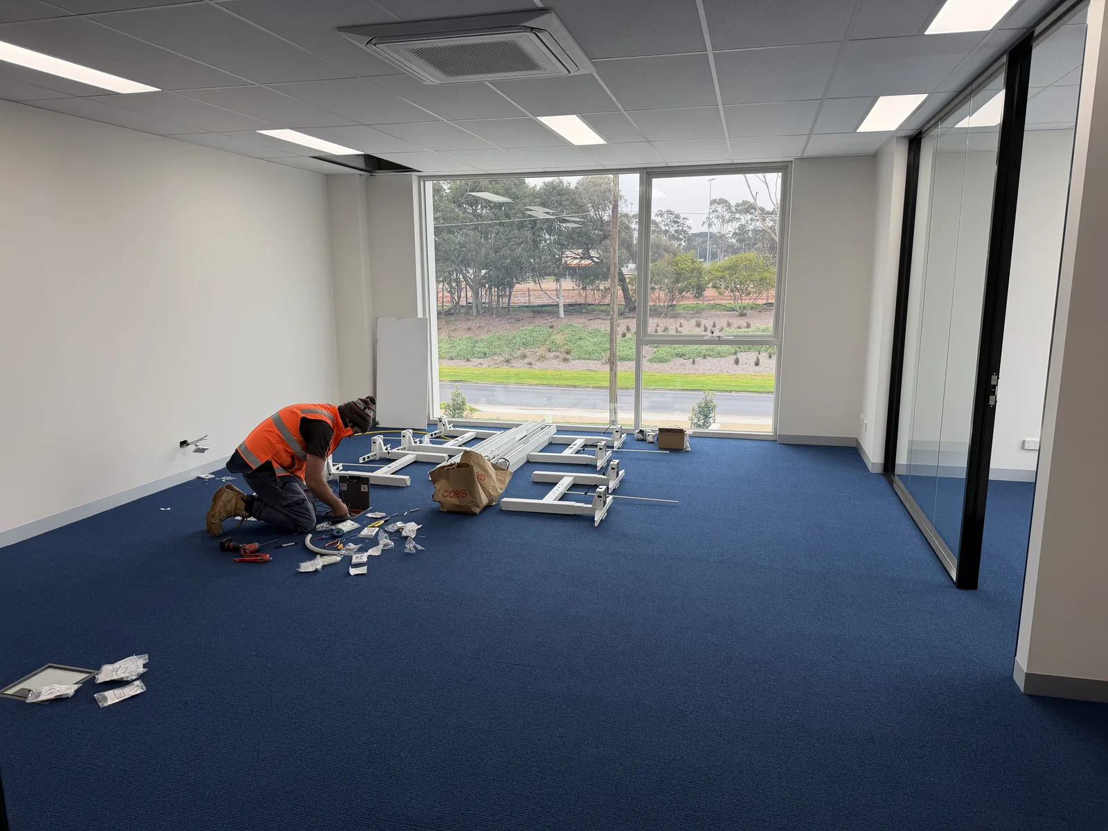 The open upstairs workspace at Free Form, Mornington during fit-out, blue carpet and a large west-facing window, a tradesperson assembling desk frames on the floor