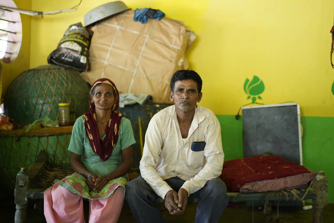 Razia and her husband Gulab sitting together at home on a traditional charpai bed, against a yellow and green wall.