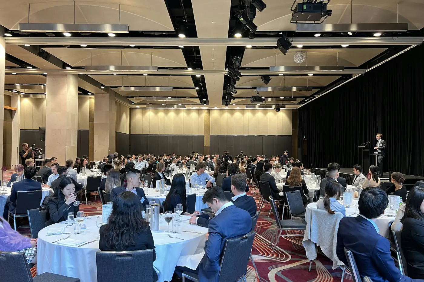 Delegates seated at round tables in a function room while a speaker addresses them from a lectern