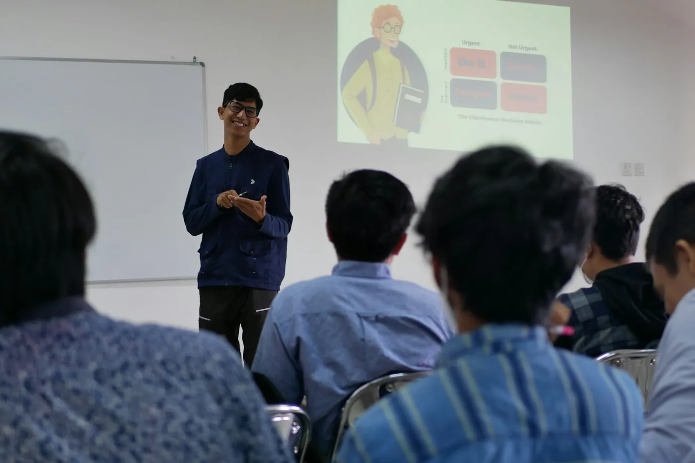 A facilitator teaching a seated group, with a slide displayed on the screen behind